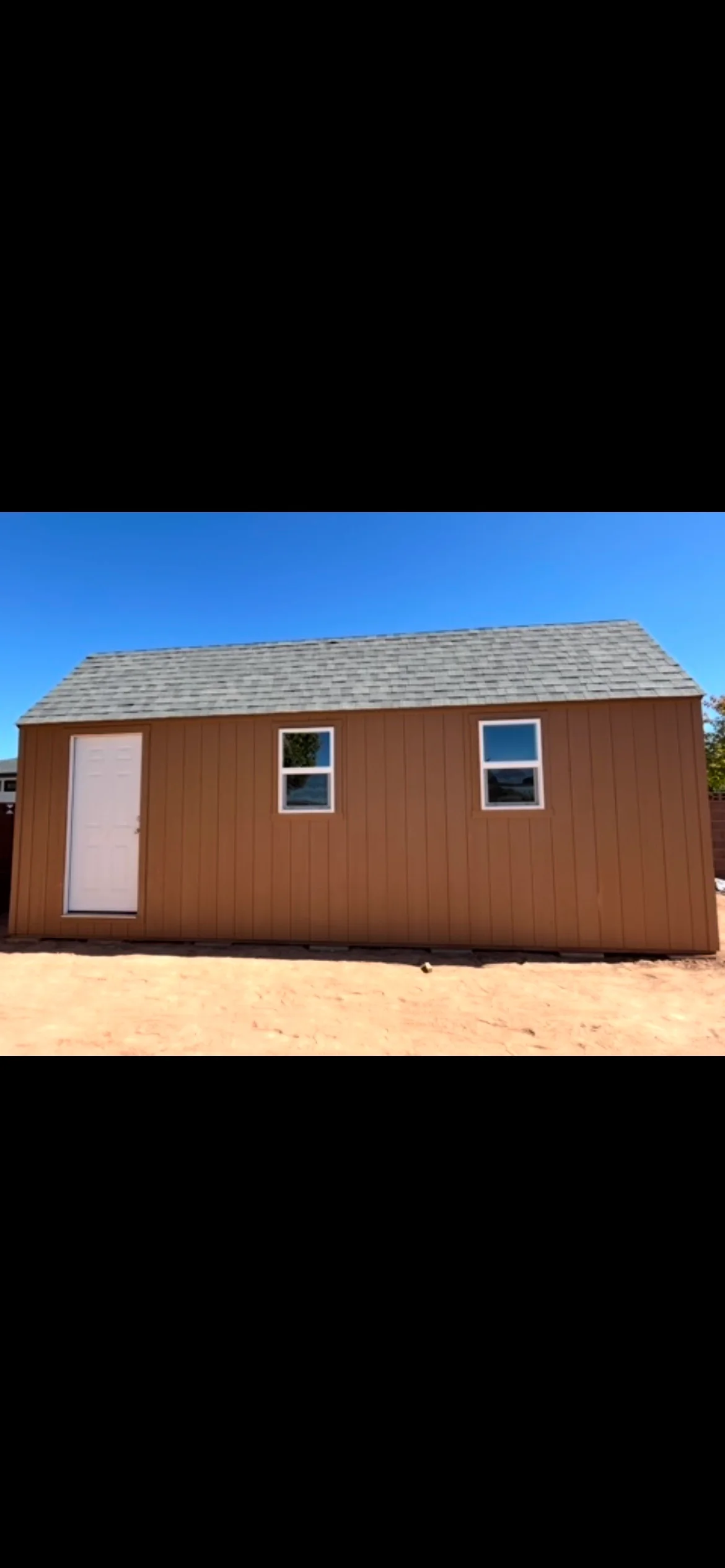 Brown barn-style storage shed with white door, two windows, and gray shingle roof on a Southern Utah lot